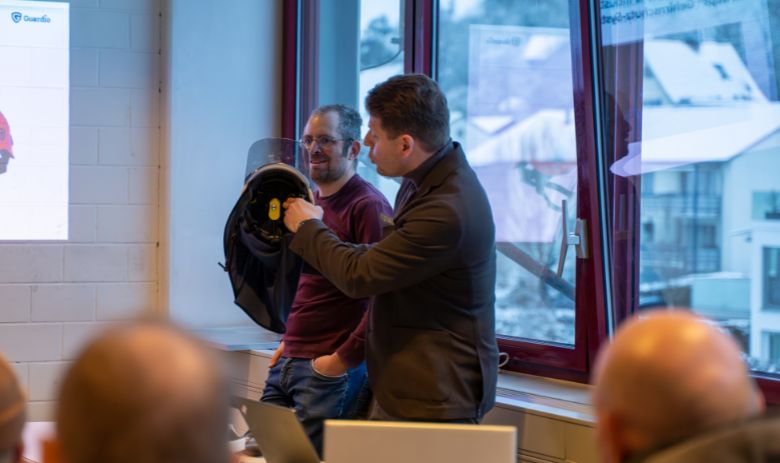 Zwei Männer stehen vor einem Publikum. Einer der beiden Männer zeigt einen Helm.