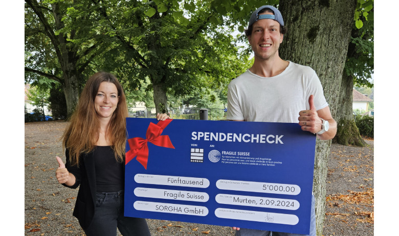 Checkübergabe Kevin Lötscher und FRAGILE Suisse Zwei Personen posieren mit einem grossen blauen Spendencheck: links Jana Bauer, rechts Kevin Lötscher. Sie lächeln und halten den Daumen hoch.