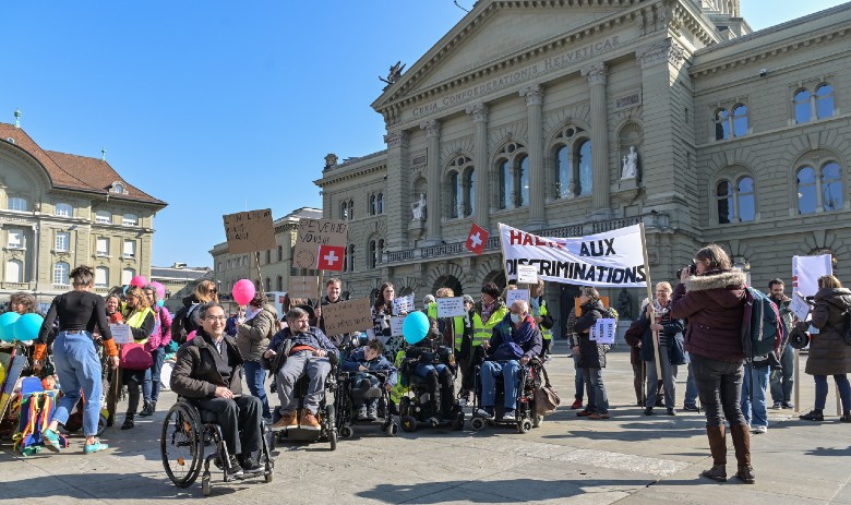 Petition Inclusion Handicap Eine Gruppe Menschen mit und ohne Beeinträchtigung ist auf einem Platz versammelt und hält Protestschilder hoch.