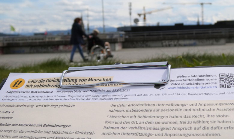Inklusions-Initiative Im Vordergrund ein Klemmbrett mit dem Unterschriftenbogen für die Inklusions-Initiative. Verschwommen im Hintergrund sind zwei Personen. Eine stösst eine andere im Rollstuhl voran.