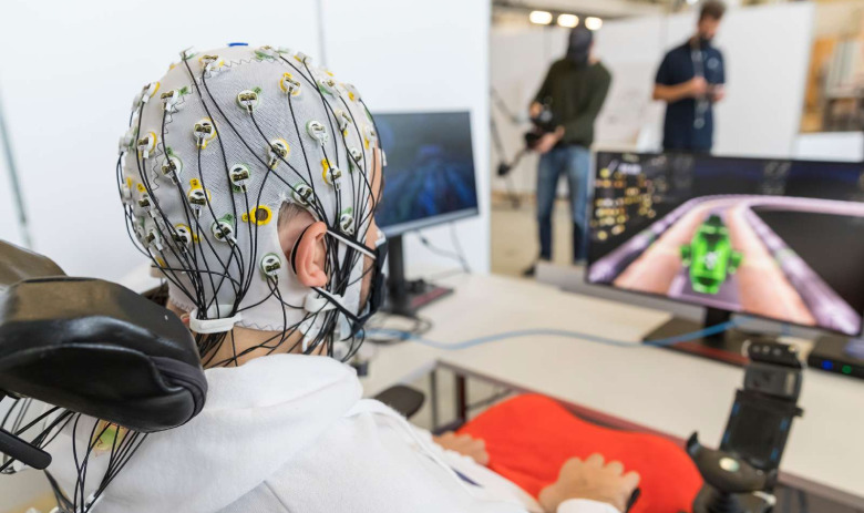 CYBATHLON Person sitzt im Rollstuhl an einem Schreibtisch mit Monitor. Auf dem Bildschirm ist eine Art Fahrzeug auf einer Rennbahn. Die Person trägt eine Haube auf dem Kopf, aus der zahlreiche Kabel hervortreten. Auf dem Schreibtisch sind keine Steuergeräte.