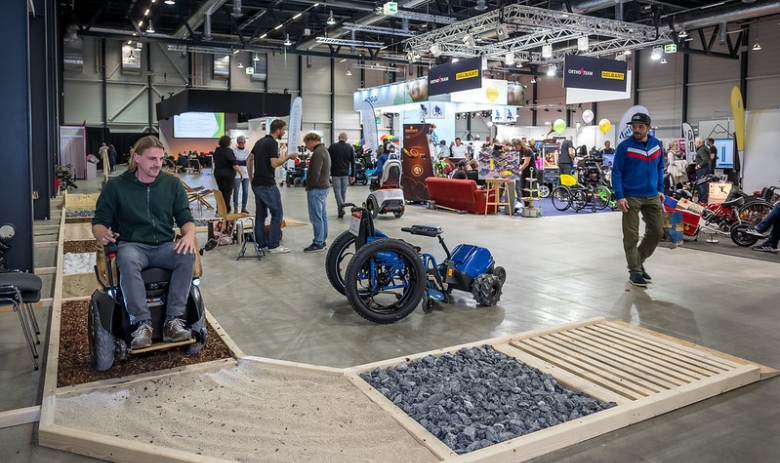 Swiss Abilities in der Messe Luzern In einer Messehalle fährt ein Mann in einem Elektrorollstuhl über verschiedene Bodenarten, wie Holzschnitzel, Sand oder Steine.