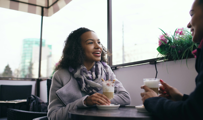 Une femme avec des cheveux bouclés tient une boisson dans sa main, elle discute avec une autre personne