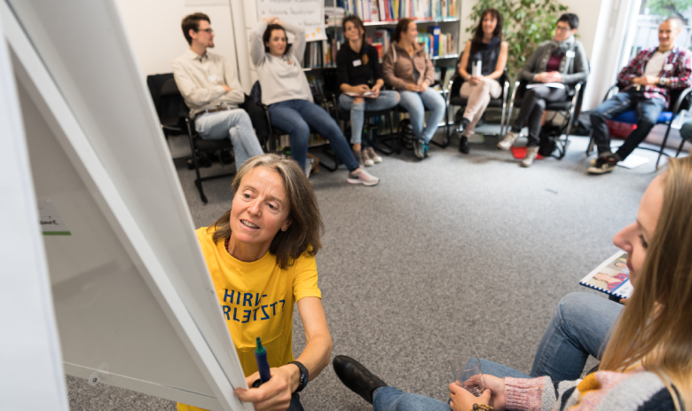 Weiterbildung Eine Frau im gelben FRAGILE-T-Shirt schreibt auf ein Flipchart. Im Hintergrund sitzen mehrere Personen in einem Halbkreis und schauen zu.